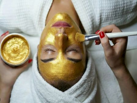 A person wearing a gold facial mask getting beauty treatment, with a brush applying product; tub of gold mask nearby, head wrapped in towel.