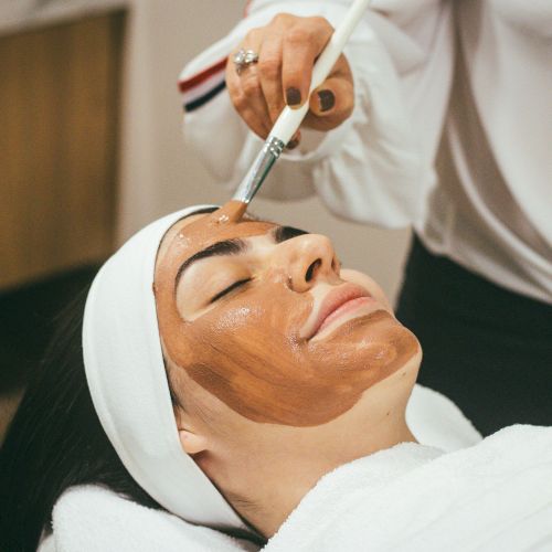 A person wearing a headband is getting a facial treatment; a practitioner applies a white cream or mask to their forehead.