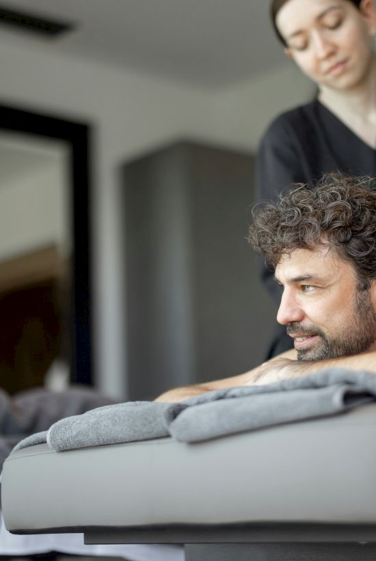 A man with curly hair lies face down on a massage table, receiving a back massage from a therapist, while others stand and watch in a spa-like room.
