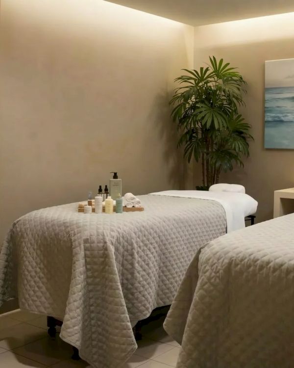 A cozy spa room with two massage tables, soft lighting, towels, and a potted plant, creating a calm, serene retreat atmosphere.