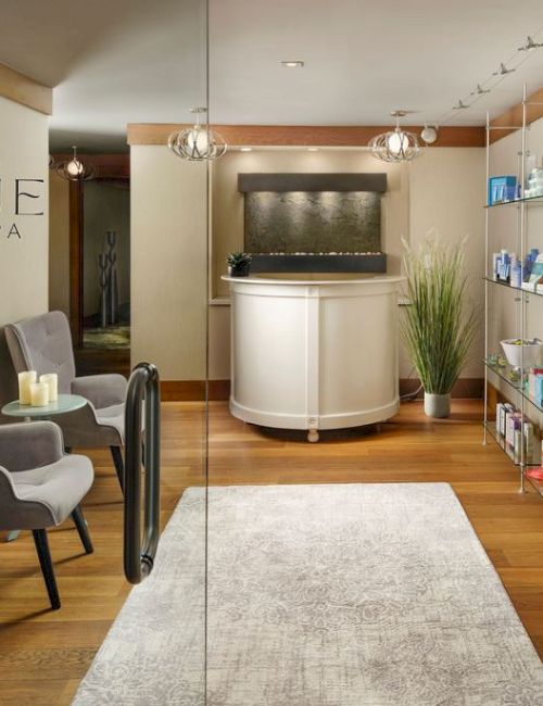 A modern spa reception with a curved white counter, seating area, glass shelves stocked with products, and a calm, inviting vibe.