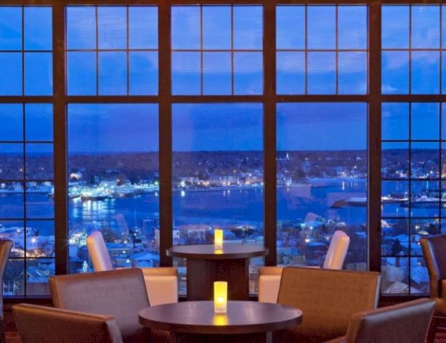 A cozy lounge with warm seating, candles on small tables, and a panoramic city view through tall windows at dusk. End.